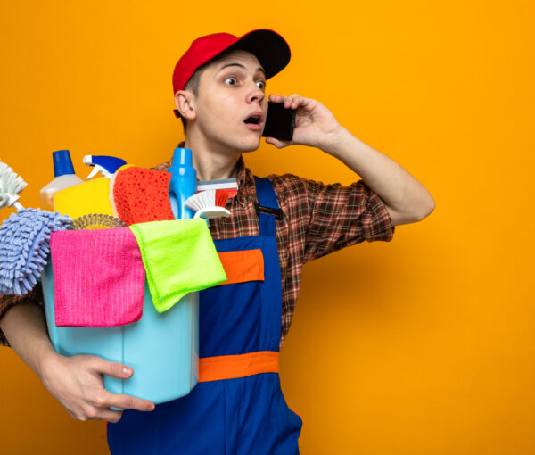 concerned young cleaning guy wearing uniform and cap holding bucket of cleaning tools speaks on phone isolated on orange background