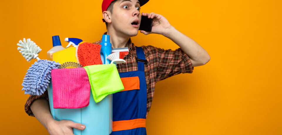 concerned young cleaning guy wearing uniform and cap holding bucket of cleaning tools speaks on phone isolated on orange background
